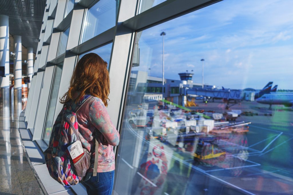 Pretty young girl traveler with a backpack next to the panoramic windows of the passenger terminal international airport watching the planes on the airfield.