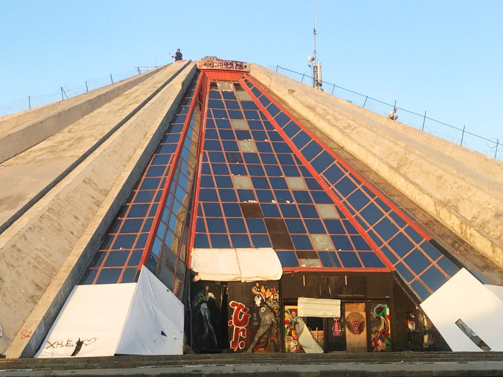 Tirana pyramid before it was renovated. You can see people stood on top and sliding down the sides of the 21 metre concrete structure. There is graffiti at the base of the pyramid .