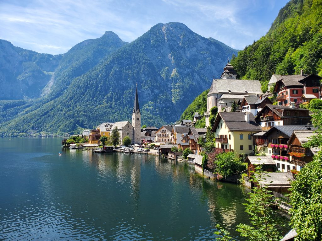 Hallstatt, Austria. The village is perched on the edge of a lake and is surrounded by tall mountains and lots of green trees. 