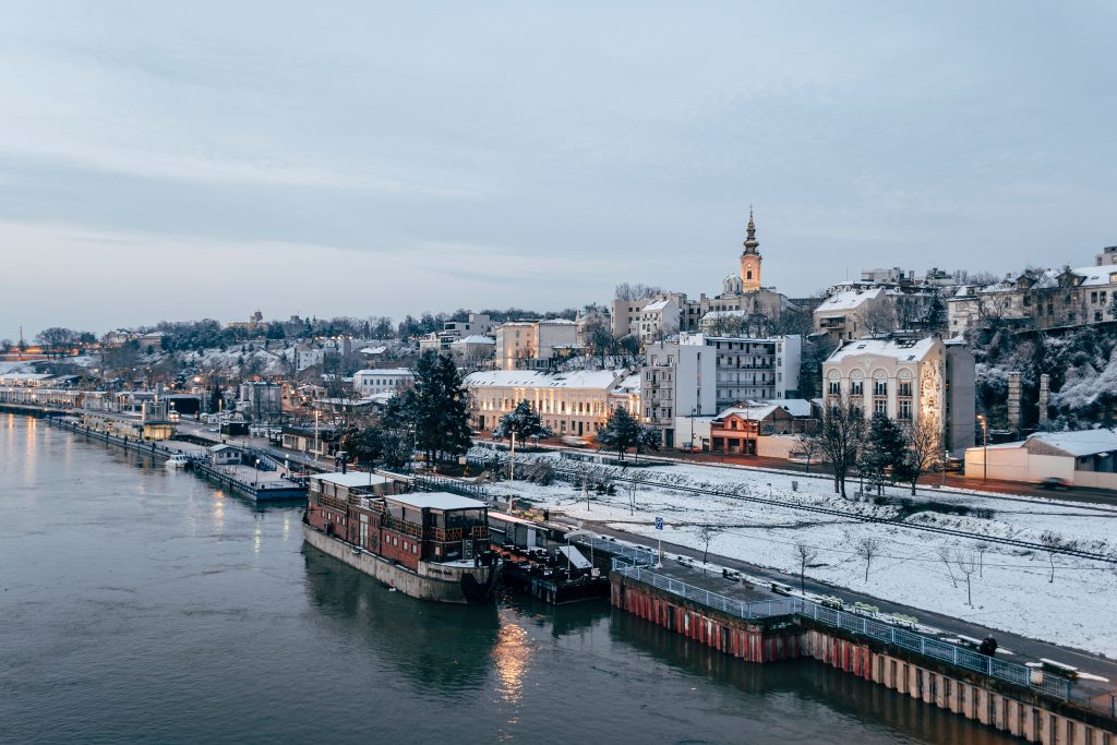 Belgrade, Serbia in winter. There is snow on the ground and on the roofs of the buildings. The river is in the foreground with some large boats moored at the riverside