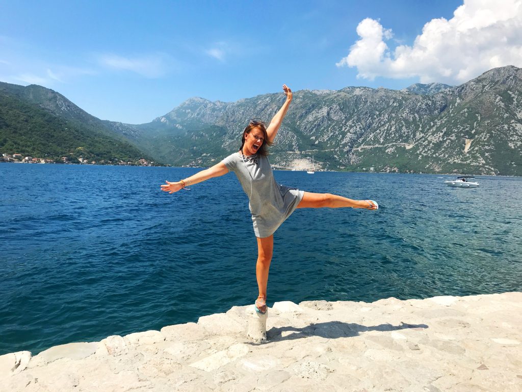 JJ jumping in front of the fjord in Kotor, Montenegro. There are mountains across the water and blue sky above. 
