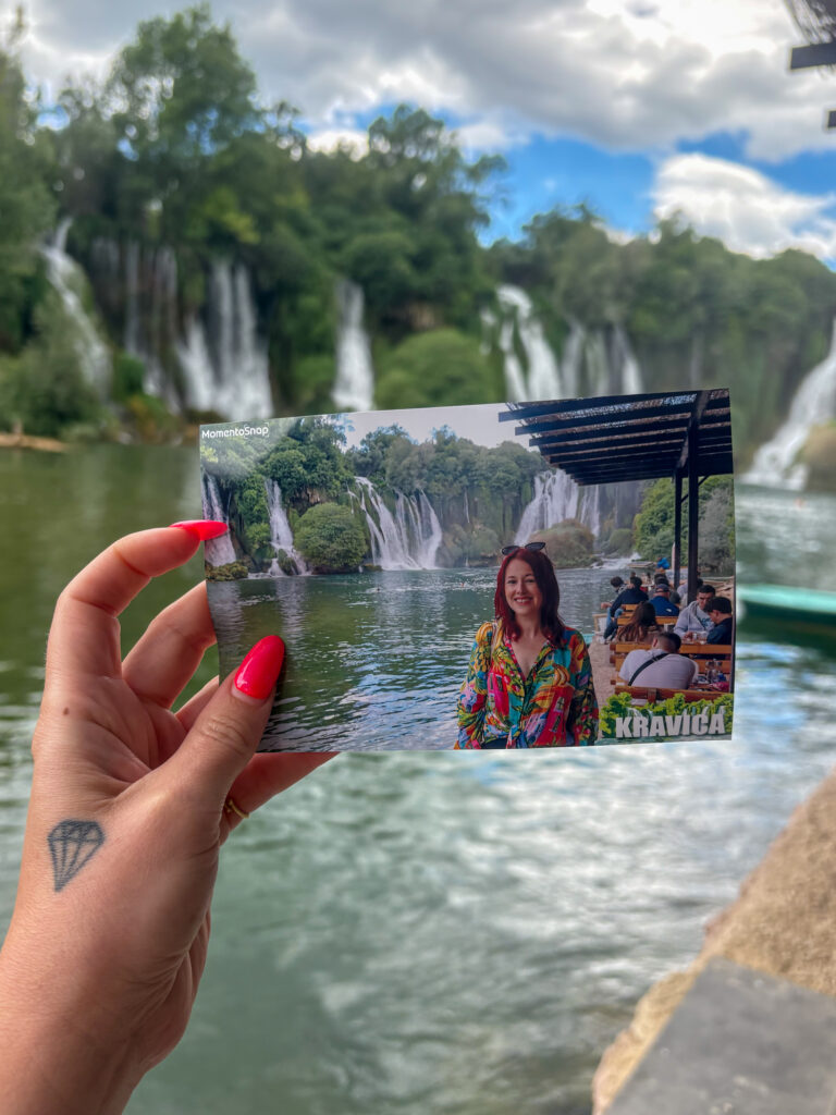 JJ's hand holding up a printed photo of herself standing in front the Kravica waterfalls. The actual waterfalls are in the background.
